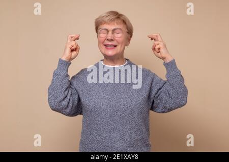 Ältere kaukasische Frau gestikuliert Finger überquert lächelnd mit Hoffnung und Augen geschlossen. Glück und abergläubisches Konzept. Stockfoto
