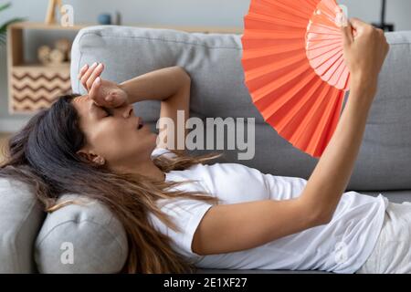 Nahaufnahme überhitzte Frau winkt Papierventilator, auf der Couch liegen Stockfoto
