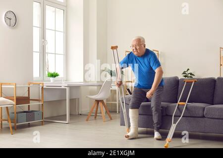 Älterer Mann mit gebrochenem Bein versucht, aufstehen Sofa und Spaziergang mit Krücken zu Hause Stockfoto