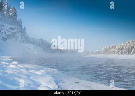 Frostiger Nebel über dem Winterfluss mit Schnee und Wald am Ufer. Erstes Eis am See an kalten Tagen. Stockfoto