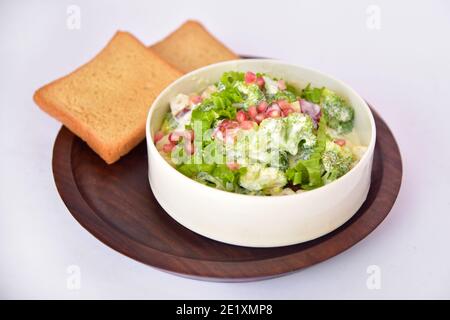 Gemüse- und Obstsalat mit braunem Brot. Salatschüssel auf Holzplatte mit Brot. apfel, Granatapfel, Brokkoli, Salat, Mayonnaise Salatschüssel. Stockfoto