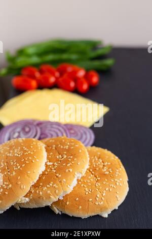 Vorbereitung für die Zubereitung eines Hamburgers, alle Zutaten: Käse, Salat, Tomaten, Zwiebeln, Brötchen liegen auf einem schwarzen Stein Hintergrund. Nahaufnahme Stockfoto