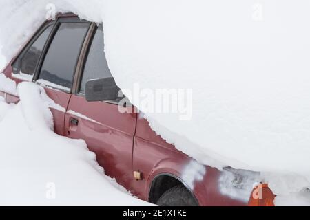 Rotes Auto unter Schneehaufen. Unerwarteter Schneefall, Gefahr auf der Straße, Schneeräumung Stockfoto