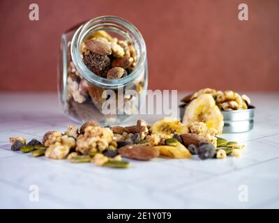 Glas voller Trail-Mix mit Nüssen, Bananenchips, Preiselbeeren und Granola auf einer marmorierten Fischgrätfliese mit einer Terrakotta-Wand dahinter. Hea Stockfoto