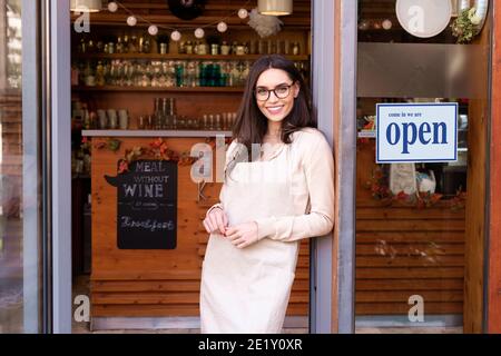 Aufnahme einer lächelnden jungen Cafe Show-Besitzerin, die mit gekreuzten Armen in der Tür stand. Schild an der Glastür öffnen. Stockfoto
