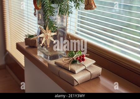 Weihnachtsgeschenke in ökologisches Recyclingpapier auf einer Fensterbank eingewickelt, mit Tannenzweigen und Strohornamenten im Hintergrund Stockfoto