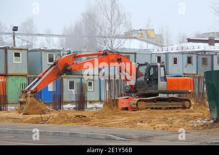 Ein großer orangefarbener Bagger auf breiten Eisenbahnen steht auf einer Baustelle auf dem Sand vor dem Hintergrund von temporären Bauerhäusern. Stockfoto