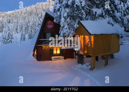 Fantastische Winterlandschaft mit Holzhaus mit Licht in den Fenstern in verschneiten Bergen und Nachthimmel. Weihnachtsurlaub und Winterurlaub Konzept Stockfoto