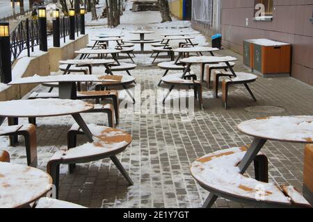 Tische und Bänke mit Schnee bedeckt stehen auf der Straße der Stadt auf einem Bürgersteig neben dem Restaurant Winter in Russland. Stockfoto