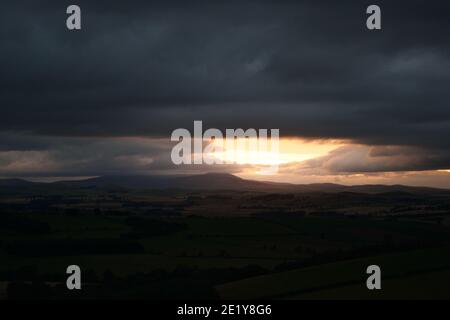 Ein Schimmer von orangefarbenem Sonnenuntergang in einem schweren, dramatischen, launischen Himmel aus grauen Sturmwolken, über einer großen flachen Ebene mit Hügeln und Bergen in der Ferne Stockfoto