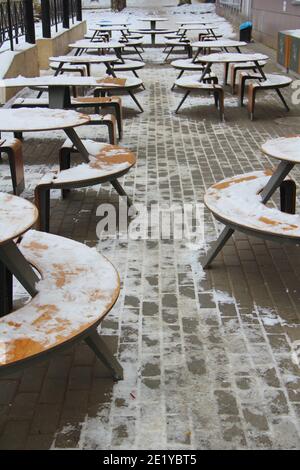 Tische und Bänke mit Schnee bedeckt stehen auf der Straße der Stadt auf einem Bürgersteig neben dem Restaurant Winter in Russland. Stockfoto