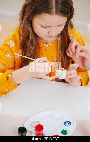 Lächelndes glückliches kleines Mädchen, das Ostereier mit Pinsel hinter einem Tisch malt Stockfoto