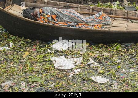 Ein Mann schläft auf einem Boot auf dem verschmutzten Buriganga Fluss in einem Wintermorgen. Dhaka, Bangladesch Stockfoto