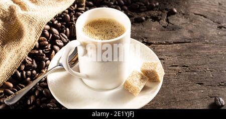 Rustikaler Hintergrund mit Tasse Kaffee Espresso in weißem Porzellan tasse mit gerösteten Kaffeebohnen auf altem Holzhintergrund mit leinentasche Stockfoto