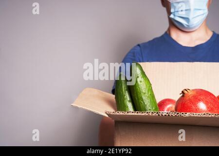 Ein weißer männlicher Kurier trägt eine Maske und trägt einen Karton mit Lebensmitteln, schnelle und sichere Lieferung Stockfoto