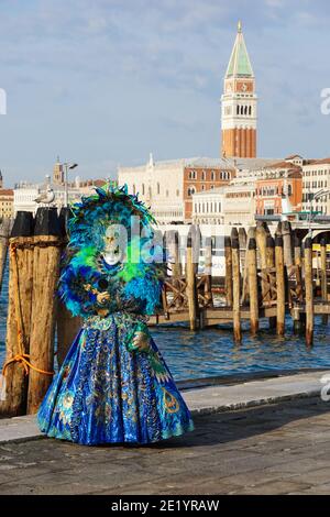 Frau gekleidet in traditionell dekoriertem Kostüm mit Federn und bemalter Maske während des Karnevals in Venedig mit dem Markusplatz Campanile hinter Venedig Italien Stockfoto