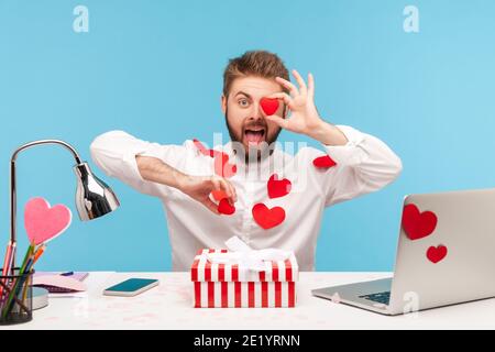 Positiv bärtiger Geschäftsmann, der ein Auge mit Spielzeug-Herz bedeckt und die Kamera mit geöffnetem Mund anschaut, am Arbeitsplatz sitzt und mit klebrigen Herzen bedeckt ist. Stockfoto