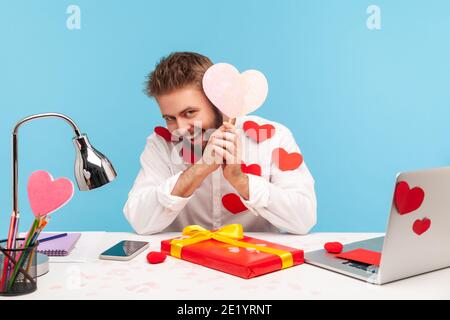 Verspielter bärtiger Mann, der am Arbeitsplatz sitzt, mit Herzstäbchen bedeckt und lächelnd, mit einer pinken Grußkarte in Herzform haltend, das Geschenk vorbereitet. Innen Stockfoto