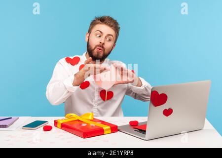 Romantischer bärtiger Mann, der Luftküsse aussenden, die über eine Webcam sprechen, mit Freundin, die am Arbeitsplatz sitzt, die alle mit klebrigen Herzen bedeckt ist, Liebe. innenstudi Stockfoto