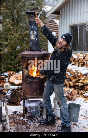 Junger Mann, der mit einer Schmiede spielt und einen Hammer hebt, lacht und vorgibt, ein Schmiedelehrling zu sein. Outdoor-Winterszene auf dem Land Stockfoto