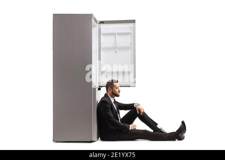 Enttäuschter Geschäftsmann, der auf dem Boden saß und sich auf einen lehnte Leerer Kühlschrank isoliert auf weißem Hintergrund Stockfoto