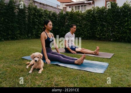 Asiatischer Mann und Frau sitzen auf Matten im Personal Pose Beim Yoga im Hinterhof in der Nähe von Hund Stockfoto