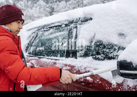 Der Mann im roten Mantel reinigt das Auto bei Schneefall mit Pinsel. Konzept für schlechtes Schneewetter. Winter schlechtes Wetter. Stockfoto