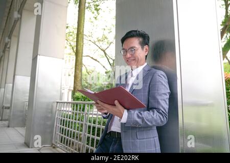 Asiatischer Geschäftsmann lehnt sich an die Säule, lächelnd, während er sein Tagebuch anschaut. Stockfoto