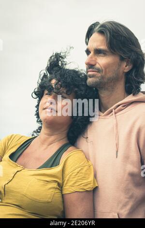 Erwartungsvolle Paare, die an bewölktem, windigem Tag in die Ferne blicken. Junge Frau, die sich auf die Brust des Freundes lehnt und einen entspannten Look hat. Vintage-Effekt Stockfoto