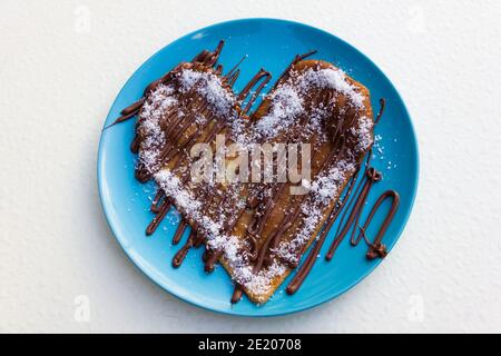 Herzförmiger Pfannkuchen mit Schokoladencreme und geriebener Kokosnuss auf blauem Teller und weißem Hintergrund. Köstliches süßes Dessert Stockfoto