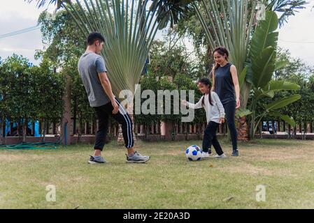 Asiatische junge Mutter, Vater und Kind Tochter Fußball spielen draußen in der Natur ein Feldgarten Park. Glückliche Familie Kind lustig spielen Fußball zusammen in s Stockfoto