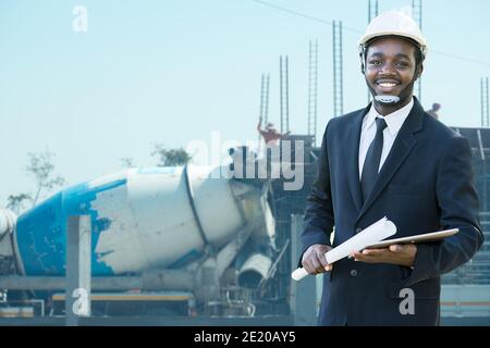 Portrait des afrikanischen Bauunternehmers Ingenieur mit Harthut halten Blaues Druckpapier und Tablett mit Gebäude im Hintergrund Stockfoto