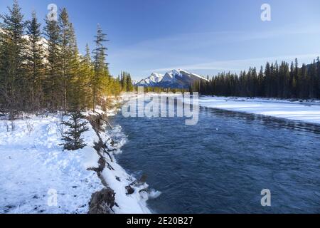 Verschneite Ufer des Bow River und ferne Rocky Mountain Peak Landschaft an einem kalten Wintertag in Canmore, Alberta in der Nähe des Banff National Park Stockfoto