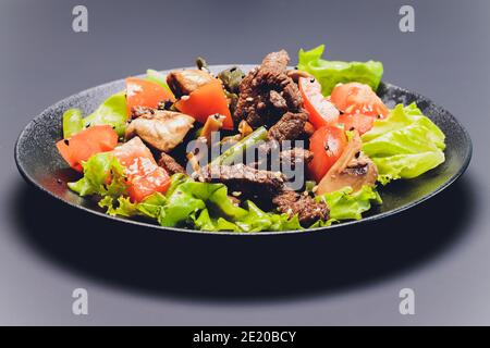 Geschnittener Rindfleisch-Tagliata-Salat mit grünen Bohnen, Kirschtomaten, frischen Rucola und Parmesan und Limettenkeilen, serviert auf einem schwarzen Teller Stockfoto