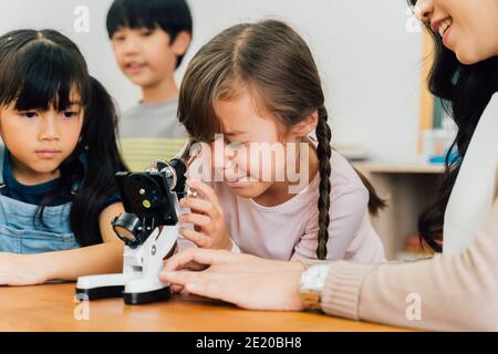 Junge Frau zeigt multiethnische Kinder Mikroskop in der Wissenschaft Klasse, Lernen, Entdeckung, Neugier. Asiatische Schule Lehrer hilft Schülern Mikroskop verwenden Stockfoto