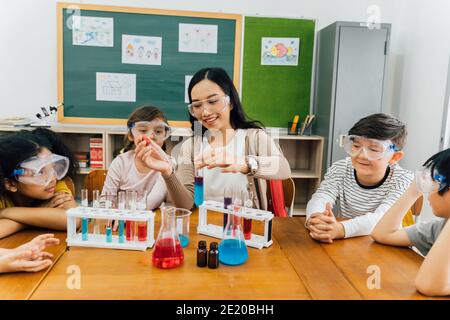 Junge Frau, die Flüssigkeiten im Reagenzglas mischt, mit multiethnischen Kindern, die sich über Wissenschaft und Chemie informieren. Asiatische Lehrer und Schüler in der Wissenschaft Klasse Stockfoto