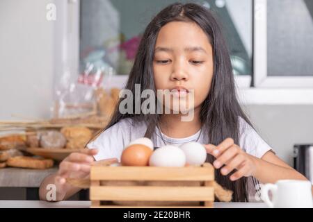 Ein kleines asiatisches Mädchen wird die Eier in einem Holzkorb auf dem weißen Kochtisch in der Küche abholen. Stockfoto