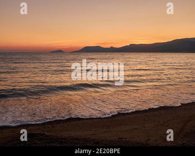 Sonnenuntergangslandschaft mit Meer und Bergen am Horizont. Küste in der Dämmerung. Am Abend surfen am Sandstrand Stockfoto