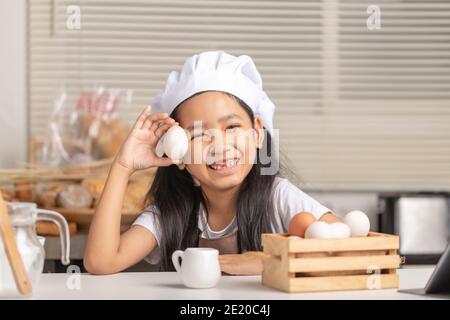 Asiatische kleine Mädchen trägt einen weißen Koch Hut hält ein Entenei und lächelt vor Glück. Eier in einem Holzkorb in der Küche. Stockfoto