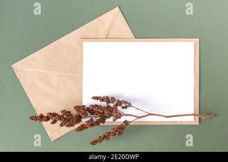Weiße leere Karte, Handwerk Umschlag und trockene Blumen Pflanzen auf grünem Hintergrund. Draufsicht Flat Lay Mockup Kopierbereich. Stockfoto
