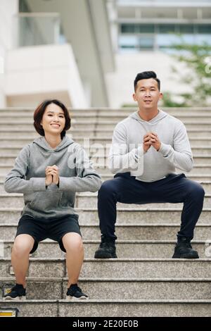 Fröhliche junge vietnamesische Paar tut Kniebeugen zusammen nach dem Laufen auf Die Straßen der Stadt am Morgen Stockfoto