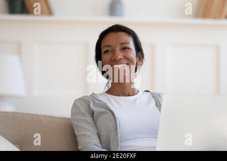 Glücklich verträumt junge afrikanische Frau sitzt auf Couch mit Laptop. Stockfoto