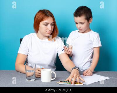 Mutter und Sohn in weißen T-Shirts zeichnen mit Aquarellen auf blauem Hintergrund. Lernen der Grundlagen des Zeichnens im Vorschulalter Stockfoto