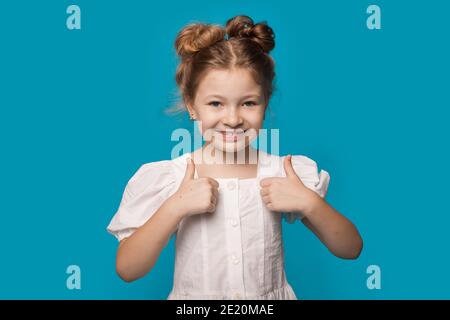 Nettes Mädchen in einem weißen Kleid ist gestikulieren die wie schild an einer blauen Wand lächelt die Kamera an Stockfoto
