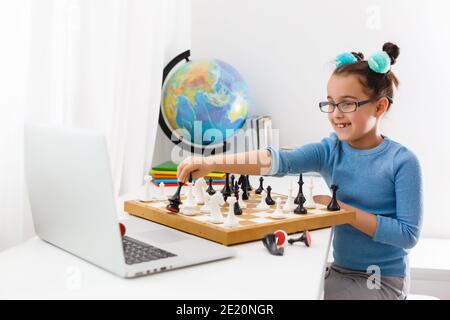 Portrait kaukasisches kleines Mädchen spielt Schach am Tisch im Innenraum mit Schachbrett. Kleines Mädchen lernt Schach auf einem Laptop online Stockfoto