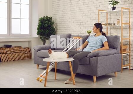 Junge Frau mit gebrochenem Bein in Gips gegossen sitzt auf Couch zu Hause und denken Stockfoto