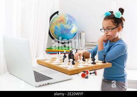 Portrait kaukasisches kleines Mädchen spielt Schach am Tisch im Innenraum mit Schachbrett. Kleines Mädchen lernt Schach auf einem Laptop online Stockfoto