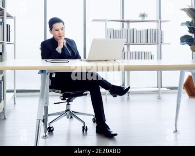 Gutaussehende asiatische Geschäftsmann in Anzug konzentrieren beobachten auf dem Bildschirm, während Arbeiten mit Laptop-Computer auf dem Schreibtisch im Büro in der Nähe von Buch Regal und Glas-Windo Stockfoto