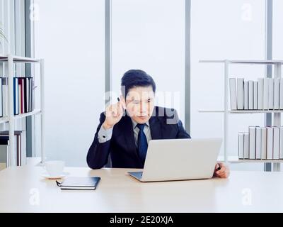 Gutaussehende asiatische Geschäftsmann in Anzug konzentrieren beobachten auf dem Bildschirm, während Arbeiten mit Laptop-Computer auf dem Schreibtisch im Büro in der Nähe von Buch Regal und Glas-Windo Stockfoto