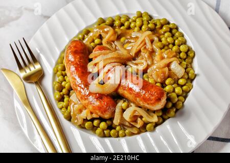 Englisches Essen auf weißem Teller: Gebratene Schweinewurst mit grünen Erbsen und klassischer Zwiebelsoße auf weißem Marmortisch mit goldenem Besteck und englischem Mu Stockfoto